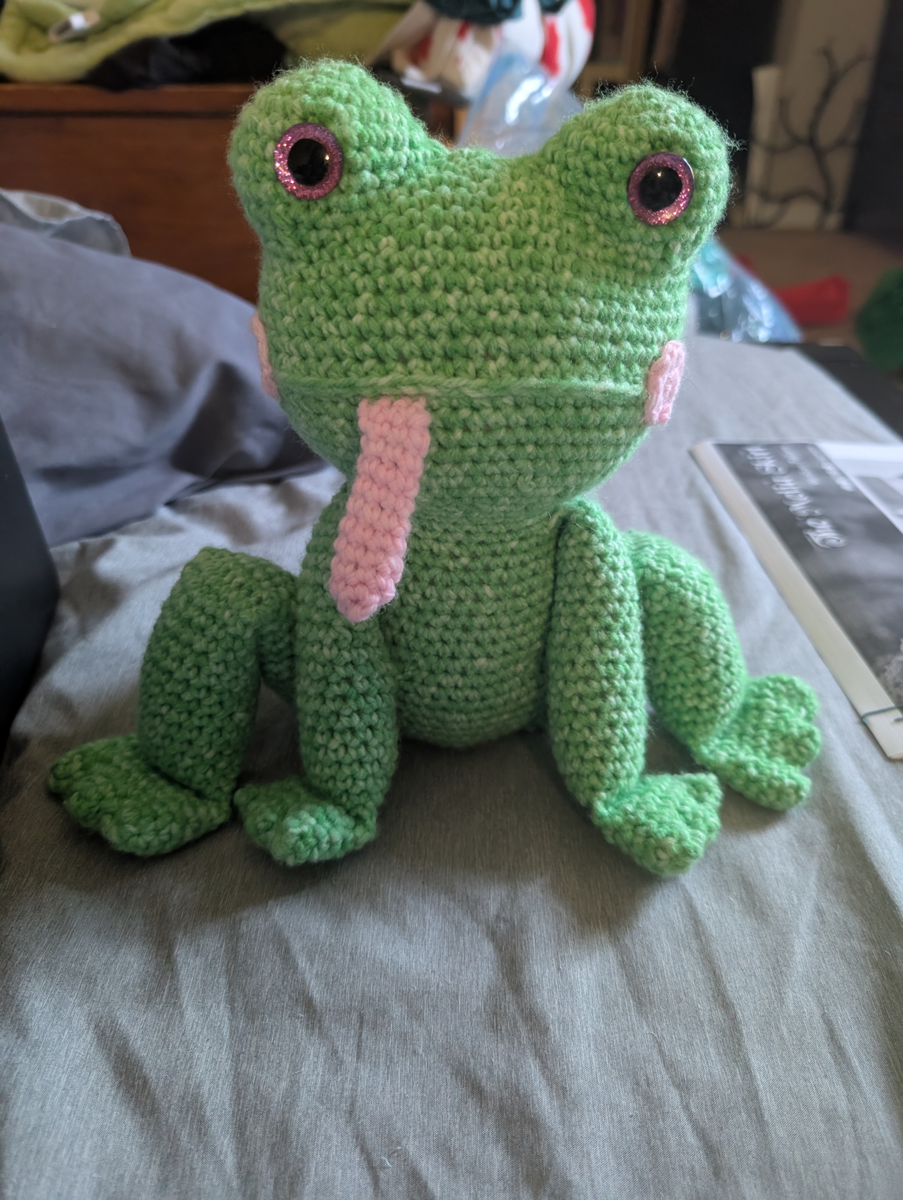 a crochet green frog with pink cheeks and a pink tongue. he has long legs bent out to each side and shorter legs in the front. it has pink glittery eyes.